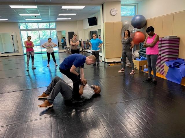 A group class in a fitness studio with two men demonstrating a self-defense technique on padded mats while others watch.